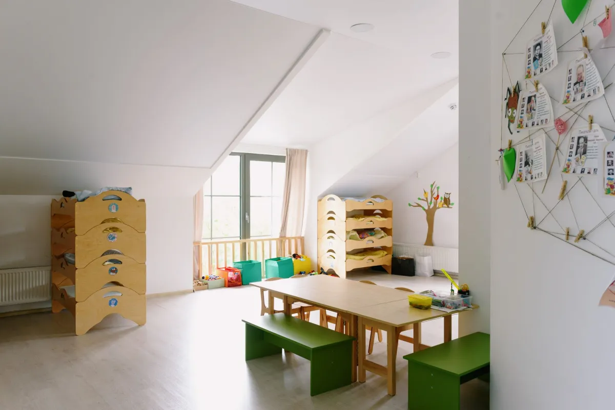 Heller Gruppenraum mit Holztischen, grünen Bänken und gestapelten Kinderbetten